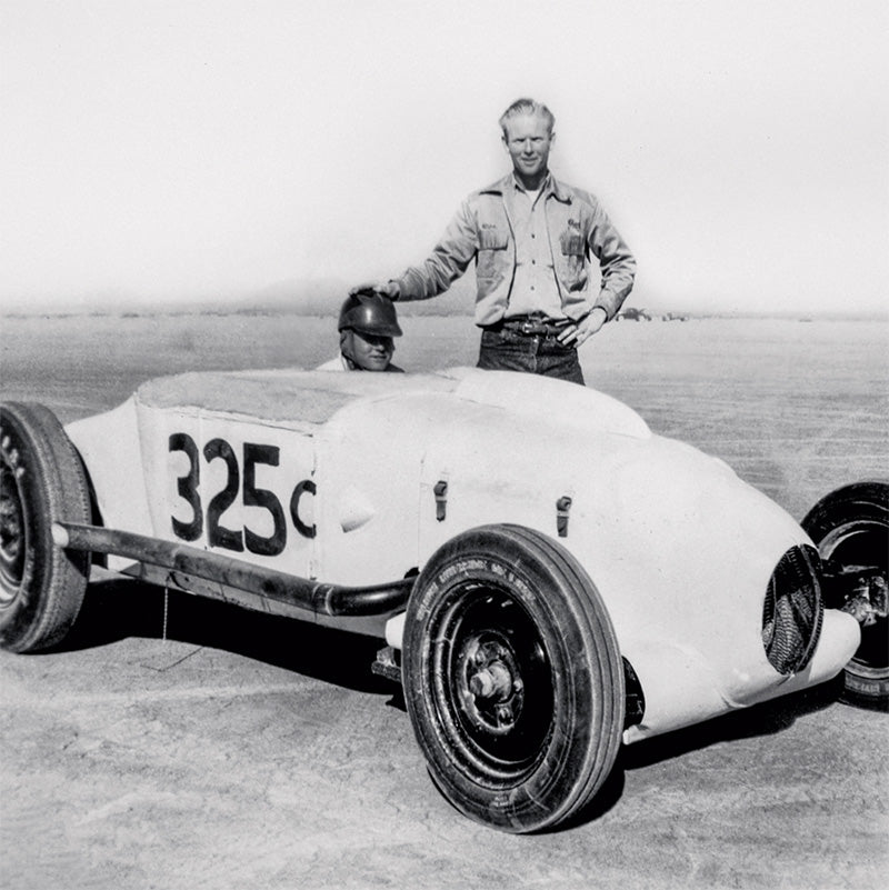 Vintage black and white photo of a race car with a driver and a person standing next to it on a track.