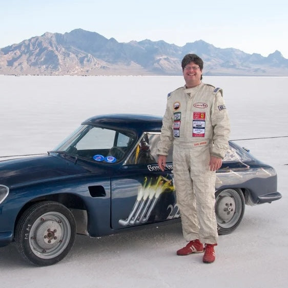 Person in a racing suit standing next to a vintage race car on a salt flat with mountains in the background