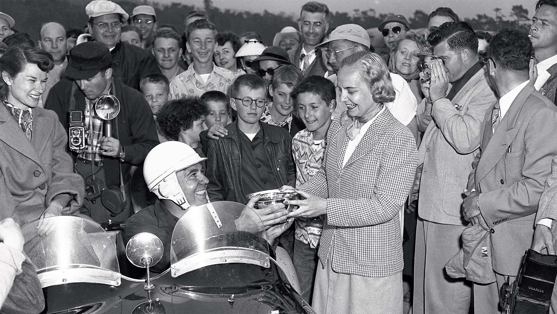 Person in a racing helmet receiving an award from a man in a suit, surrounded by onlookers.