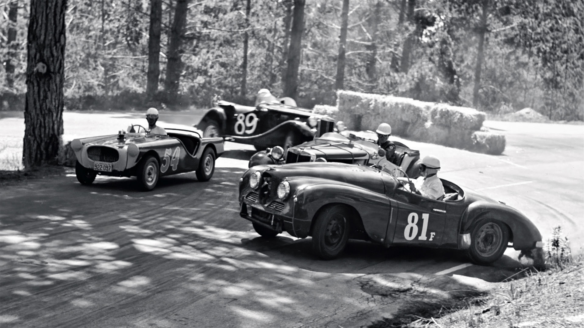 Vintage race cars on a dirt track with trees in the background