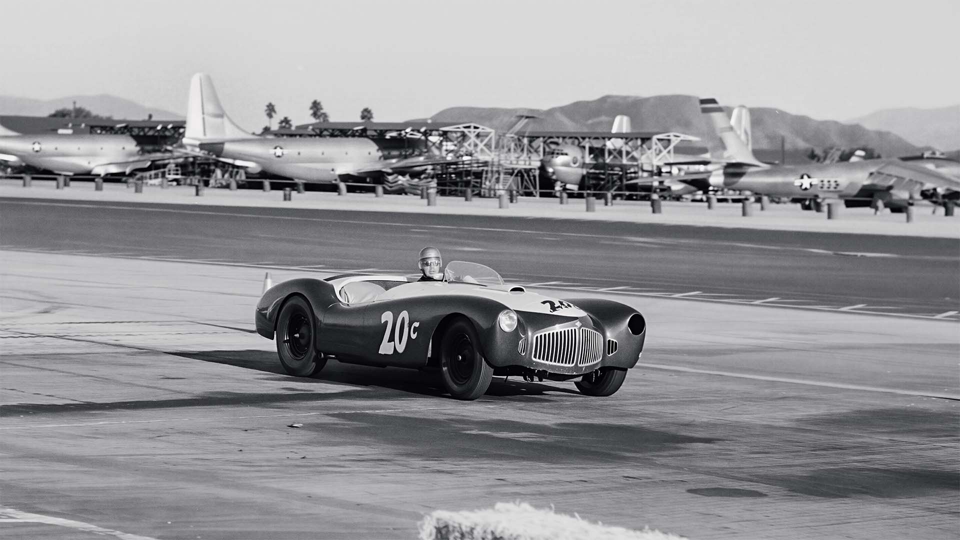 Vintage race car on an airstrip with airplanes in the background