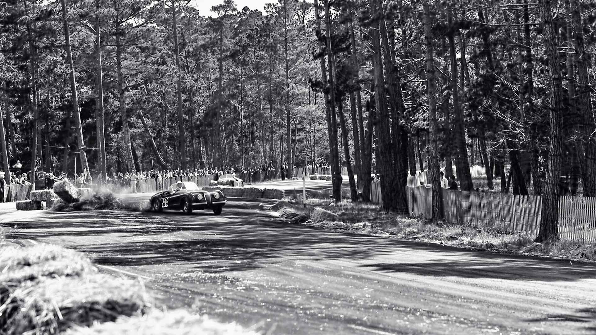 Vintage black and white photograph of a race car on a track with trees in the background