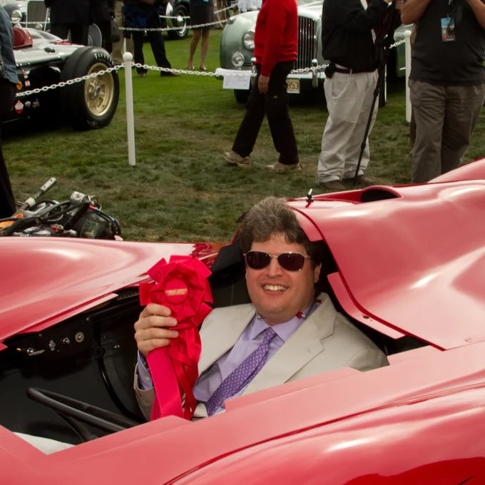 Man sitting in a red car holding a pink flower at an outdoor event with other people and cars in the background.